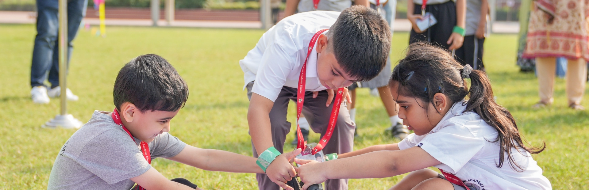  Young students at the Aga Khan Academy Dhaka participate in an outdoor teamwork activity, sitting on mats and holding each other’s feet while stretching together.